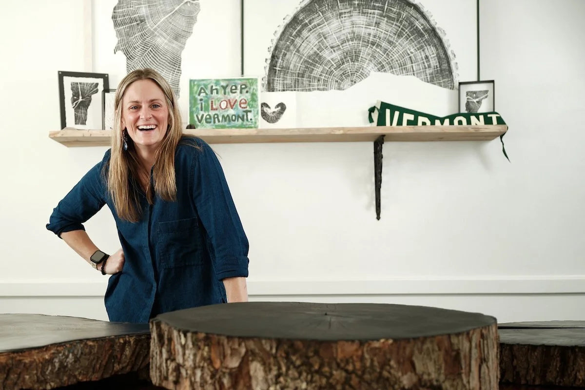 A woman with blonde hair wearing a dark blue button-up shirt stands smiling in a room with a white wall, a wooden shelf with ink prints of tree rings, and a large rustic tree-stump table in the foreground.