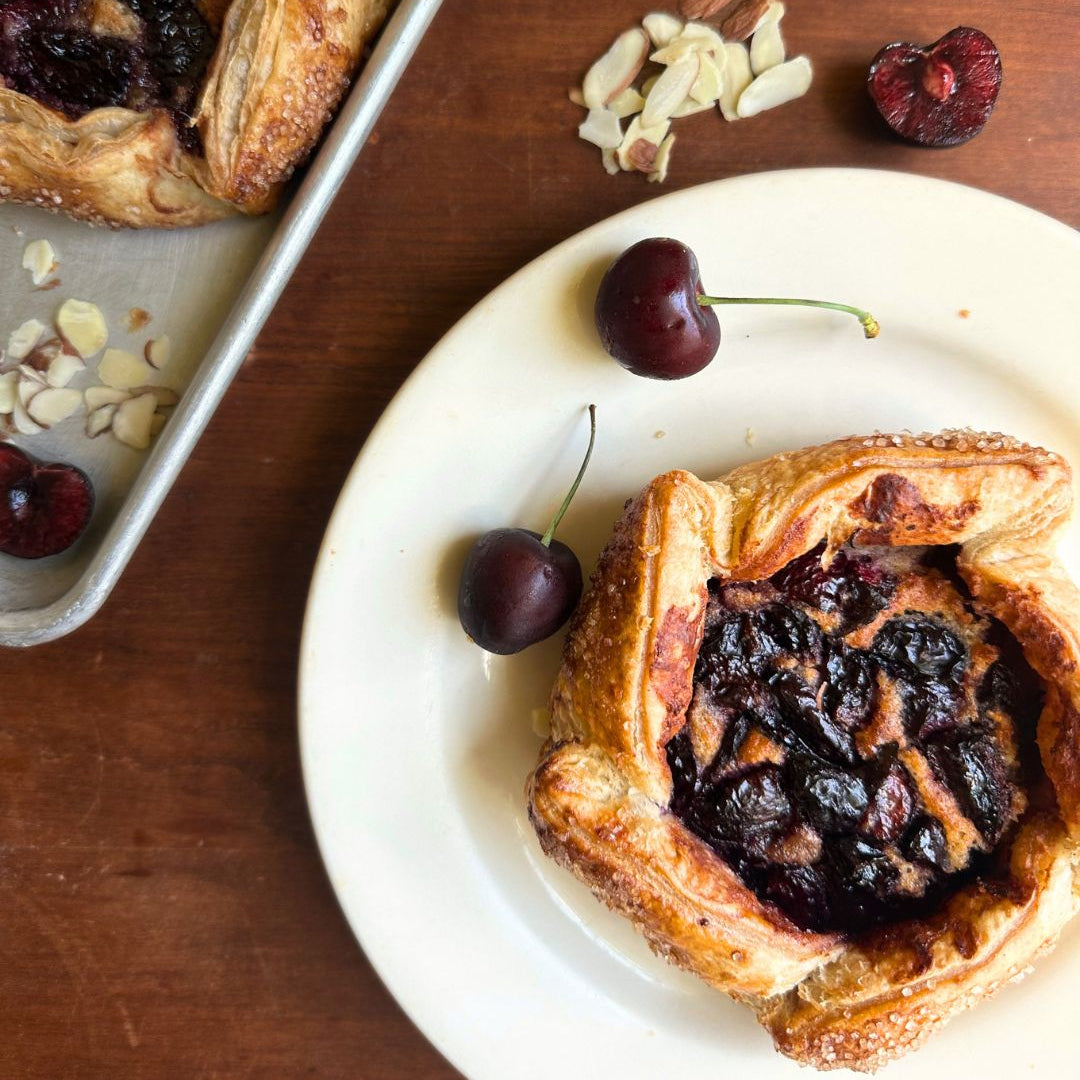 Two cherry and dried fruit pastry tartlets are shown on a wooden table, with one on a white plate and the other in a metal baking pan. Sliced almonds and whole cherries are scattered around as styling props.
