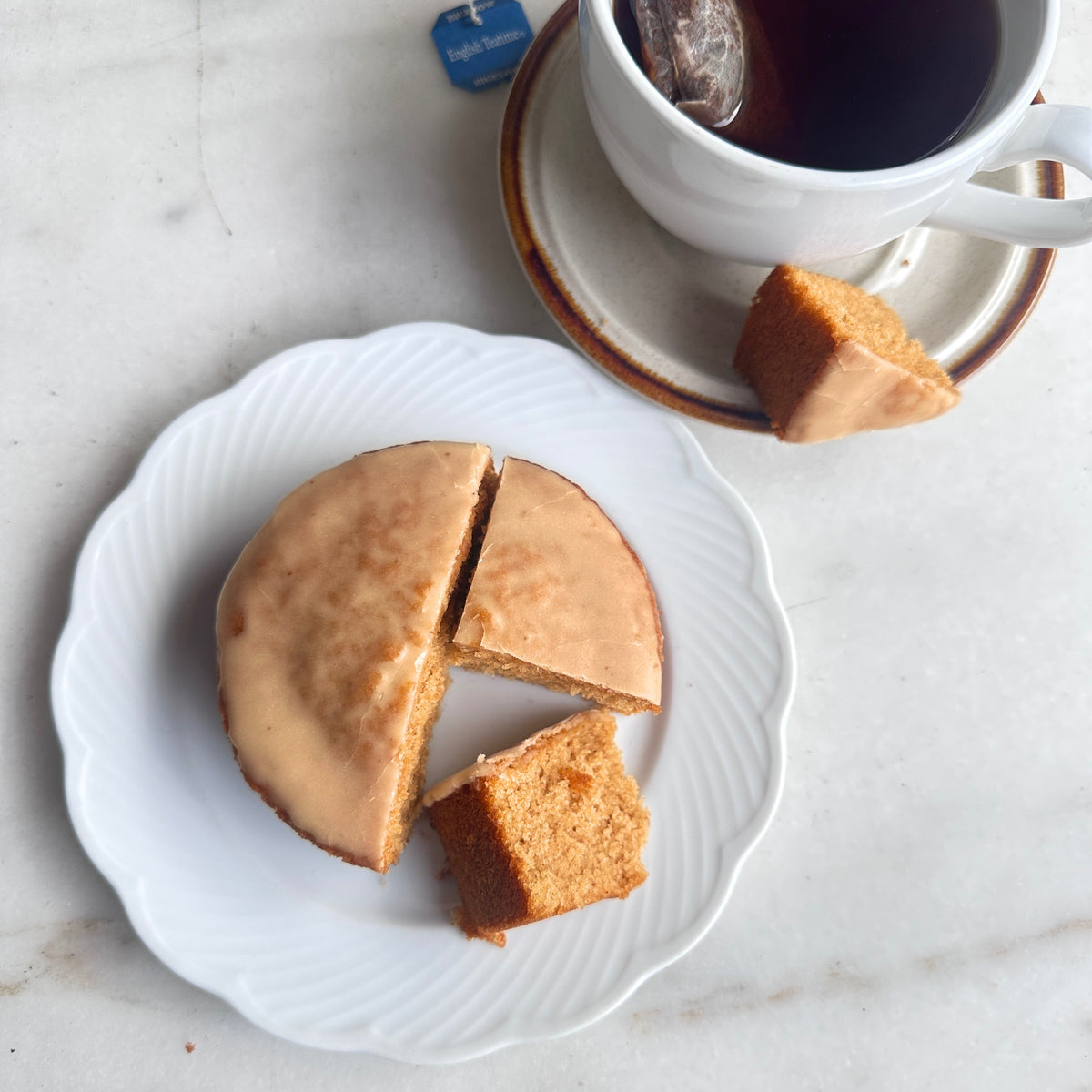 A small round cake or pastry on a white plate with a slice cut out, next to a white cup of tea on a saucer with a tea bag tag visible, all on a light marble-like surface.