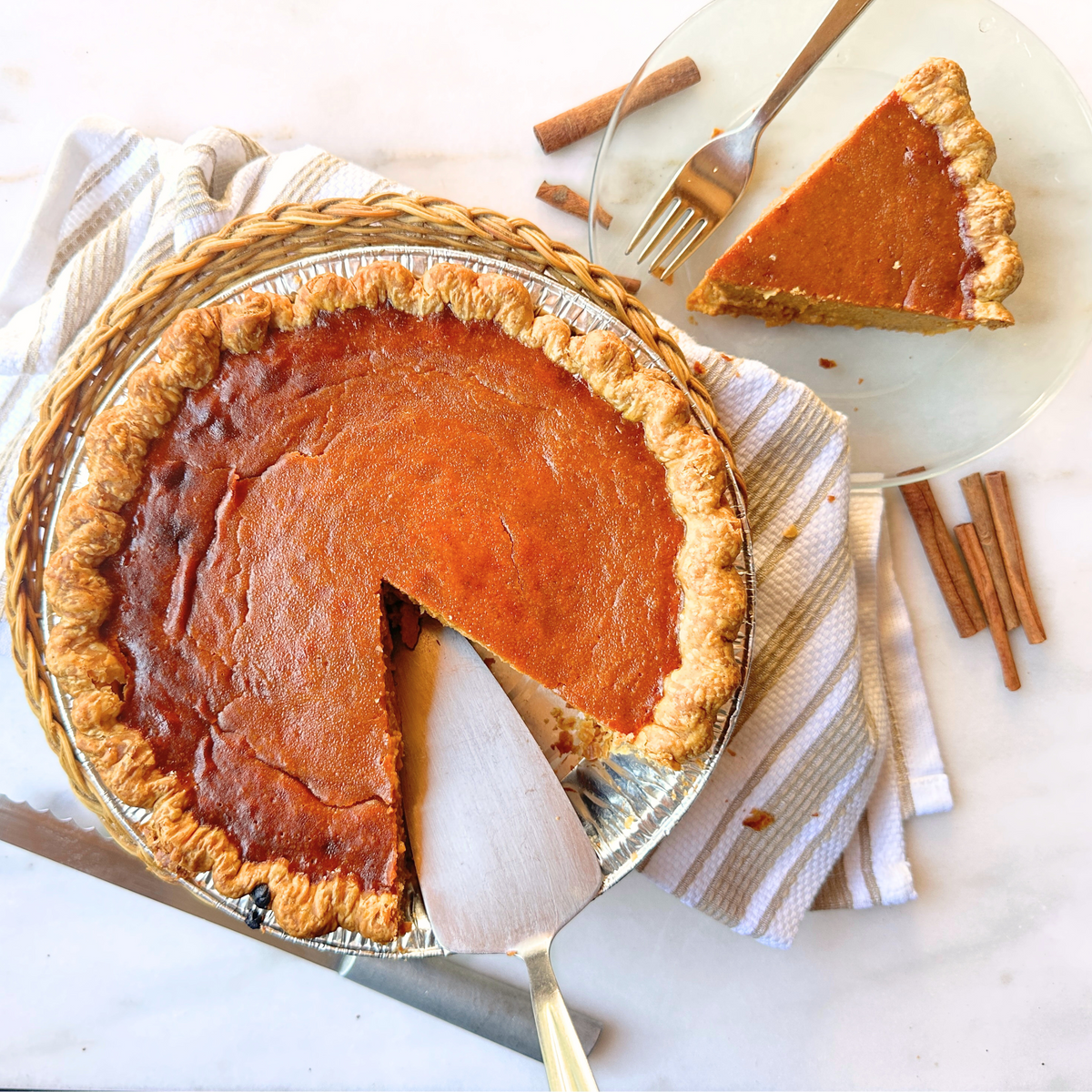 Pumpkin pie with a slice removed, on a marble surface with cinnamon sticks.