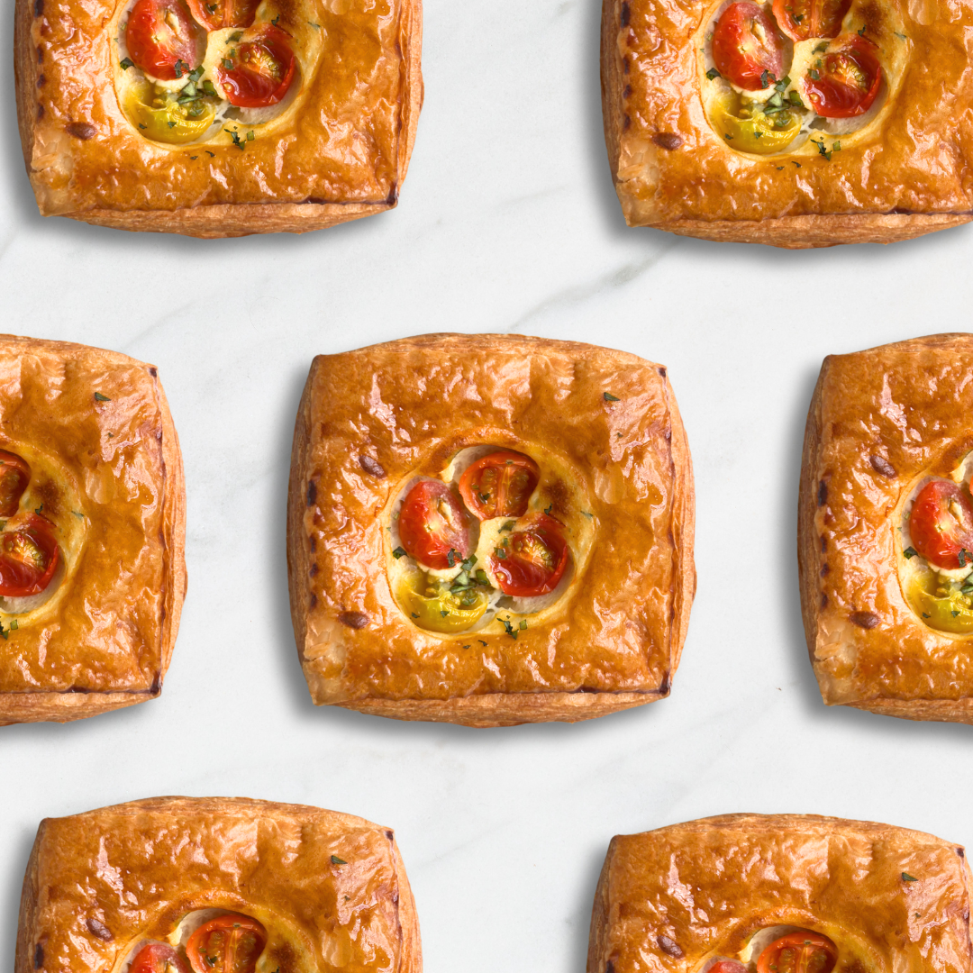 Six golden-brown baked pastries arranged in a grid on a light marble surface. Each pastry has a flaky crust and a circular topping filled with sliced red tomatoes, melted cheese, and small green herb pieces.